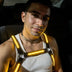 LED Harness by Neon Cowboys, an amber glowing light up harness worn over a white tank top with a choker at night, this neon led harness for men works as go-go dancer, drag, cabaret and rave performer stagewear for festivals, EDM concerts, Coachella, Pride and club nights, Neon Cowboys