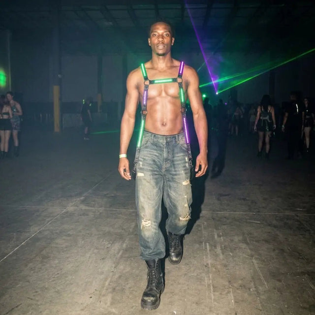 LED Harness by Neon Cowboys, a shirtless man in a green and purple glowing light up harness walking through laser beams at a warehouse rave, this neon harness and led harness for men is bold rave performer and go-go dancer stagewear for festivals, EDM concerts, EDC, Pride and club nights, Neon Cowboys