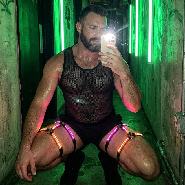 Man crouching in a green-lit hallway taking a mirror selfie in a mesh top and pink-glowing LED thigh harnesses, a color-changing led harness men leg set and neon harness stagewear for go-go dancers and rave performers at clubs and festivals, Neon Cowboys