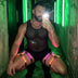 Man crouching in a green-lit hallway taking a mirror selfie in a mesh top and pink-glowing LED thigh harnesses, a color-changing led harness men leg set and neon harness stagewear for go-go dancers and rave performers at clubs and festivals, Neon Cowboys