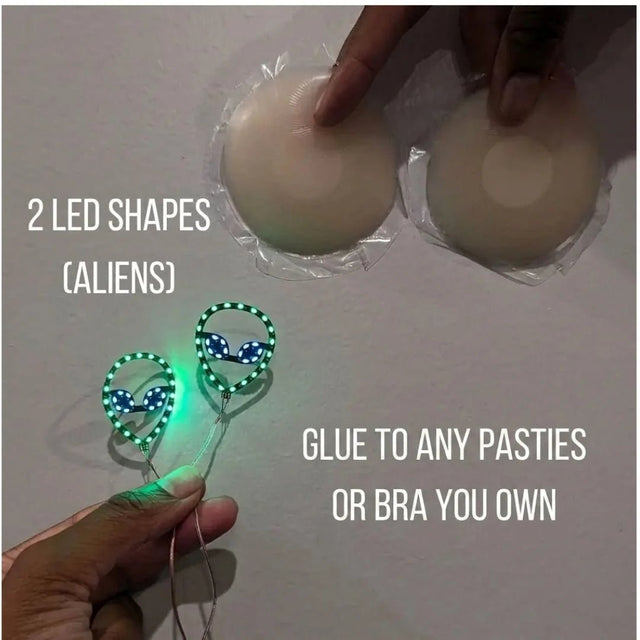 Two glowing green LED alien shapes next to silicone pasties — light-up alien nipple covers you glue onto any pasties or bra