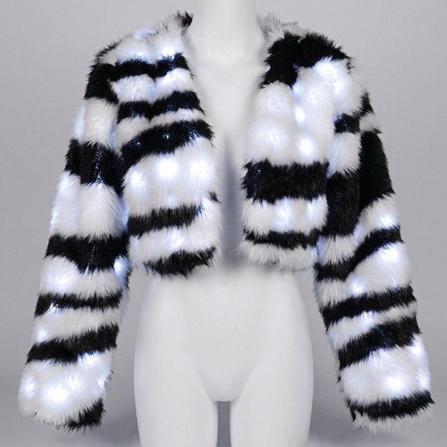 Woman dancing at a festival in a white glowing zebra LED faux fur crop jacket with silver shorts and boots, a color-changing light up coat and led costume for festival dancers and showgirl stagewear at raves, Coachella and Pride, Neon Cowboys