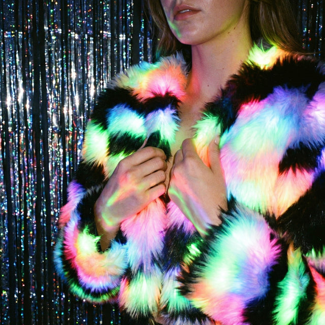 Close-up of a woman clutching a pastel rainbow color-changing LED faux fur crop jacket by a glitter curtain, a glowing light up rave coat and led costume for go-go dancers and showgirl performance outfits at Coachella, Pride and EDM concerts, Neon Cowboys