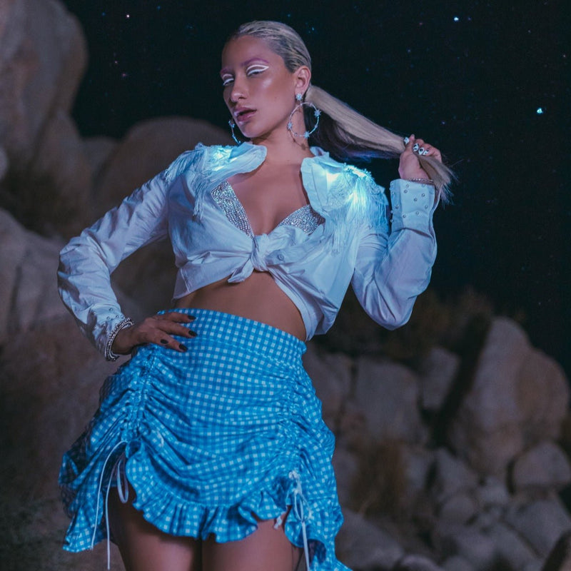 Woman at night in a white tied light-up fringe button down glowing blue with a gingham skirt, an illuminated LED fringe yoke western shirt and light up jacket-style stagewear for festival dancers and go-go, perfect for country music festivals, rodeos and western parties, Neon Cowboys