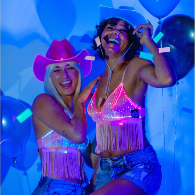 Two women in coordinating pink and blue light up fringe bralettes with cowboy hats showing matching festival outfits for country concerts and rodeo events