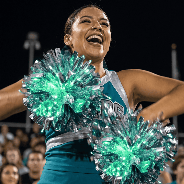 Cheerleader at a Friday night football game holding two glowing green and silver LED light-up pom poms in her team colors — remote-controlled color-changing cheerleading pom poms for high school, college, all-star and dance teams ordering custom team colors in bulk.
