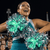 Cheerleader at a Friday night football game holding two glowing green and silver LED light-up pom poms in her team colors — remote-controlled color-changing cheerleading pom poms for high school, college, all-star and dance teams ordering custom team colors in bulk.