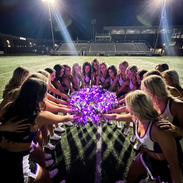 College cheer and dance squad huddled on a stadium field at night around glowing purple LED light-up pom poms — bulk custom team pom poms for cheerleading squads, dance teams, drill teams and pom lines in matching team colors.