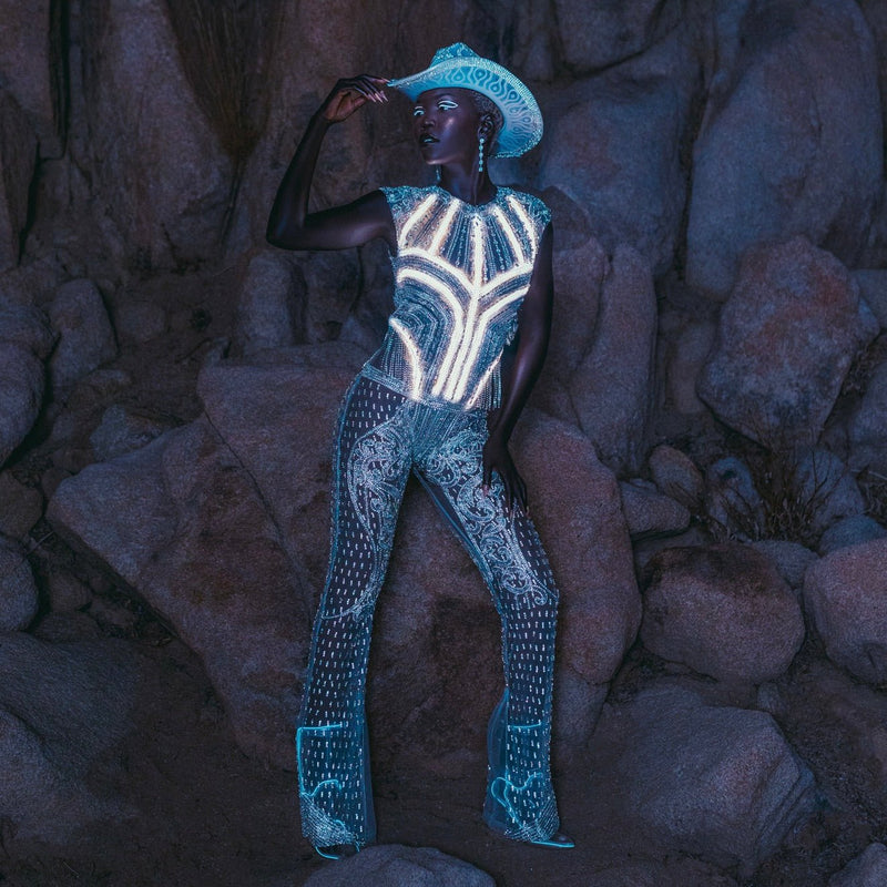 Model poses on rocks in a Neon Cowboys light up rhinestone bodysuit glowing white with a matching cowboy hat, led bodysuit rave outfit stagewear for go-go dancers, burlesque performers and showgirls at raves, EDM concerts and music festivals, Neon Cowboys