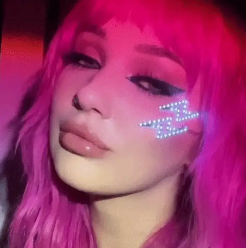 A pink-haired woman wears white-glowing LED lightning bolt face stickers under her eye at a party, LED face jewelry and face gems for festival makeup, stage makeup, and rave outfits with a rocker chic look, perfect for concerts, EDM festivals, and Coachella, Neon Cowboys lightning face stickers, Neon Cowboys