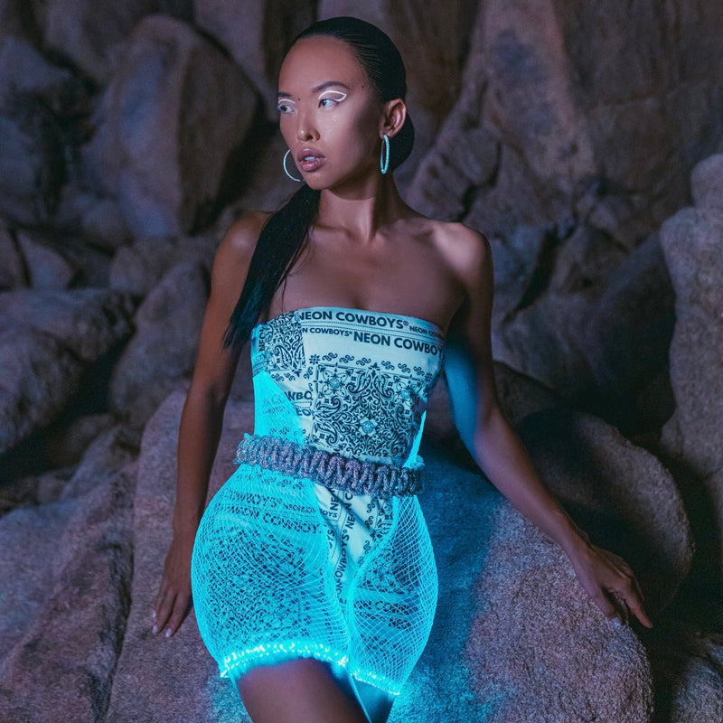 Model in a Neon Cowboys mini bandana net dress glowing teal with a logo bandana bodice on desert rocks, led net dress stagewear for go-go dancers, burlesque performers and showgirls, stunning glow for festival fashion, raves and nighttime events, Neon Cowboys