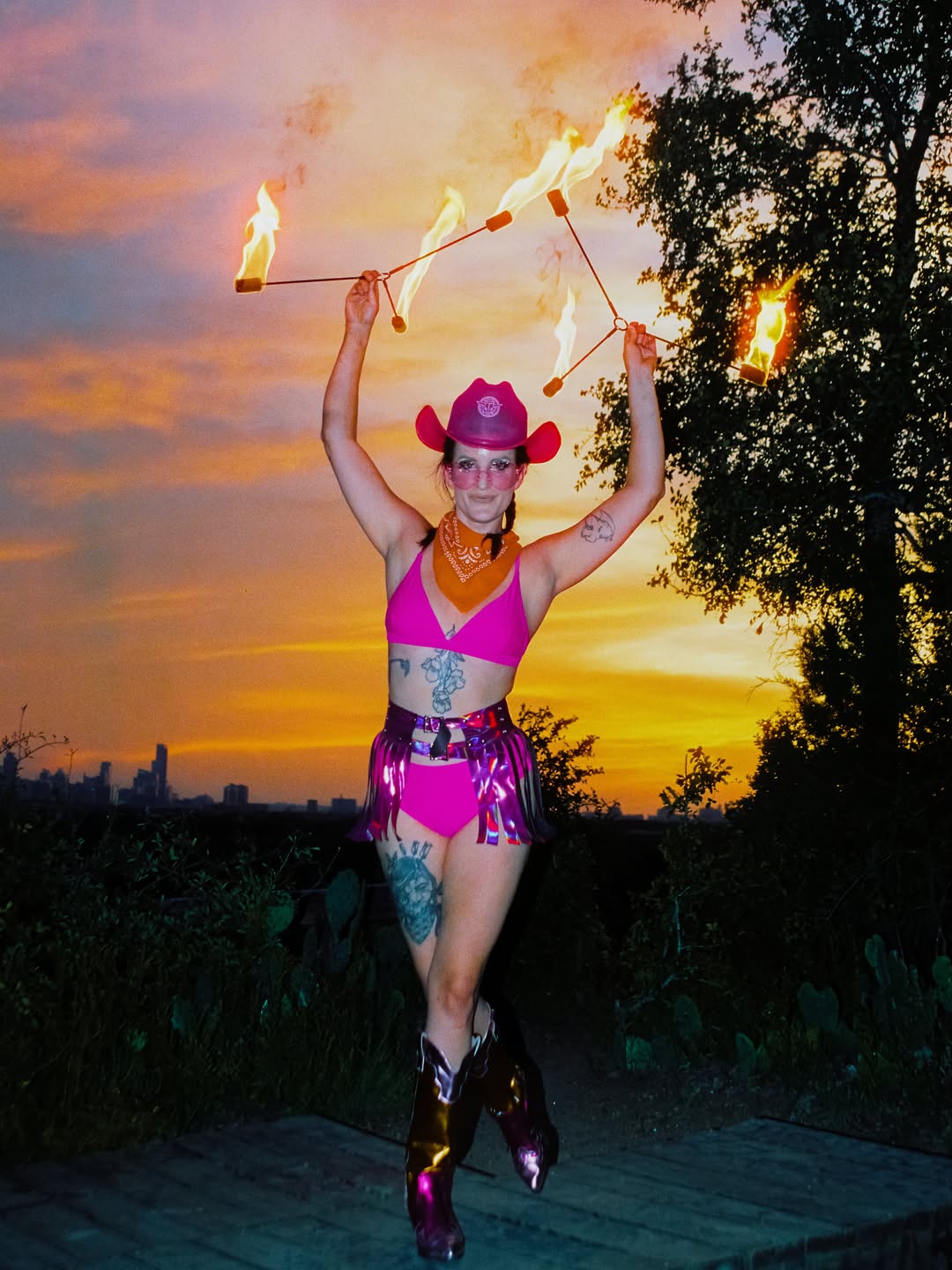 Fire performer in Neon Cowboys pink LED cowboy hat, holographic fringe skirt and chrome boots spinning fire at sunset in Austin Texas, LED and light-up stagewear for performers