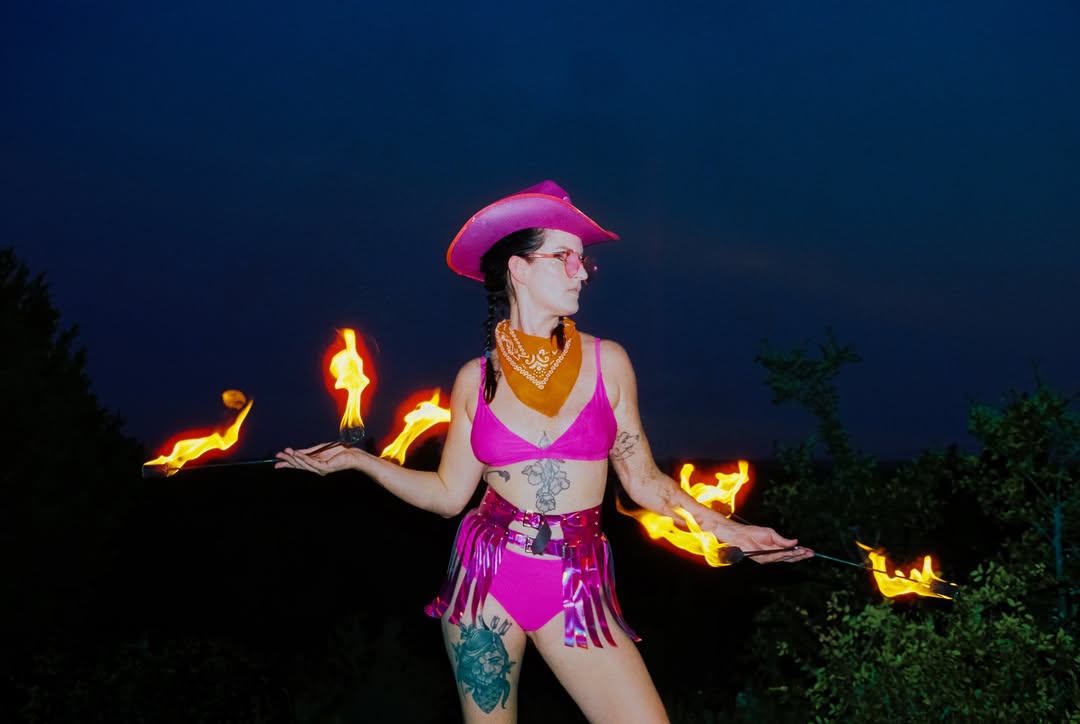 Fire performer in Neon Cowboys pink LED cowboy hat, holographic fringe skirt and chrome boots spinning fire at sunset in Austin Texas, LED and light-up stagewear for performers