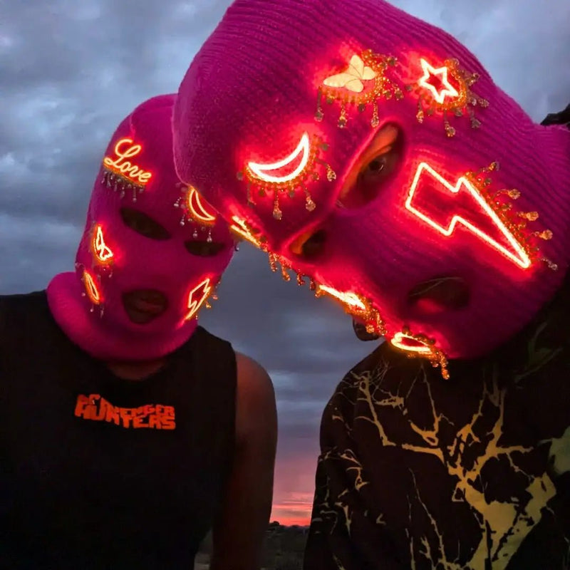 Two people at dusk in pink LED light up patched balaclavas glowing with moon and lightning designs, illuminated neon ski masks and light up face masks for EDM festivals, cyberpunk fashion, rave outfits, and winter nightlife events, Neon Cowboys neon balaclava, Neon Cowboys