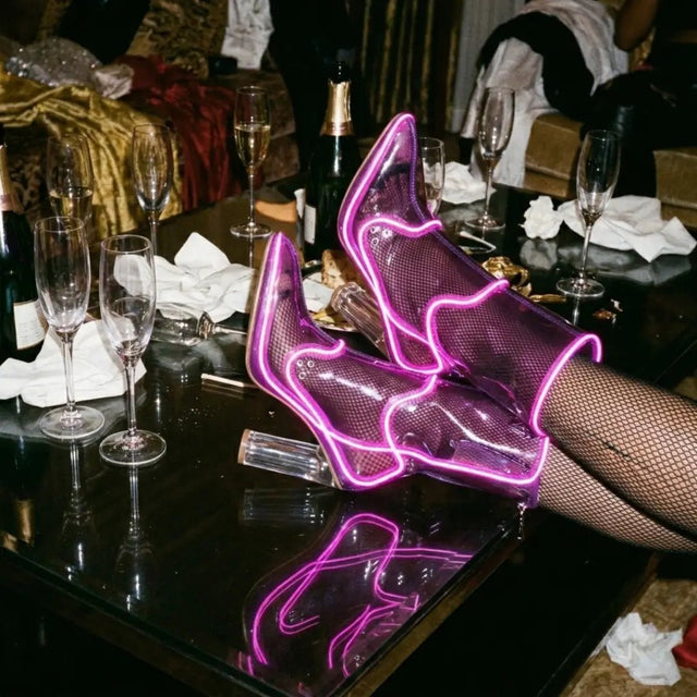 Legs in fishnets and purple glowing LED light up boots resting on a table with champagne glasses, these Out Past Midnight light up heels and light up cowgirl boots are made for raves, nightclub nights and bachelorette parties, Neon Cowboys