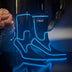 A pair of black boots glowing blue beside pink carnations on a reflective surface, these Out Past Midnight light up cowboy boots and light up heels are made for raves, music festivals and bachelorette nights, Neon Cowboys