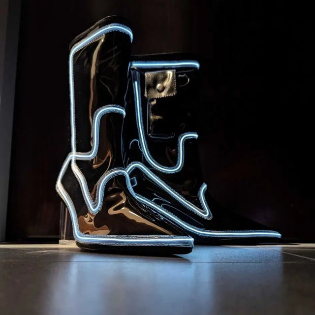A pair of black patent light up boots outlined in white and blue glow on a wood floor, these Out Past Midnight light up cowboy boots and light up heels are made for raves, music festivals and nightclub nights, Neon Cowboys