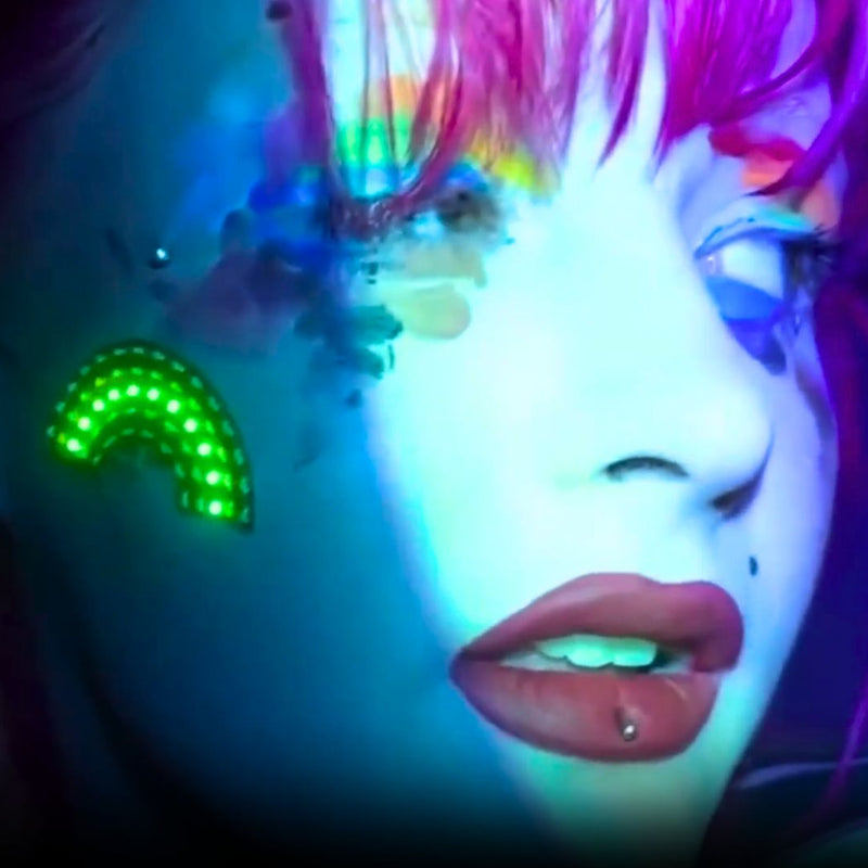 Close-up of a face under UV light with a glowing green rainbow LED face sticker on the cheek, led face stickers and led face jewelry rainbow face gems for Pride, festival makeup and rave makeup, perfect for Pride parades, music festivals, raves and drag shows, Neon Cowboys