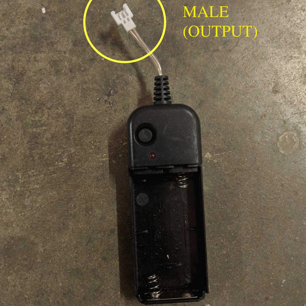 Black battery pack on concrete with a yellow circle highlighting the male output connector, a Neon Cowboys LED replacement inverter pack for light up apparel and led costumes, a handy spare power source for festivals, raves and all-night events, Neon Cowboys
