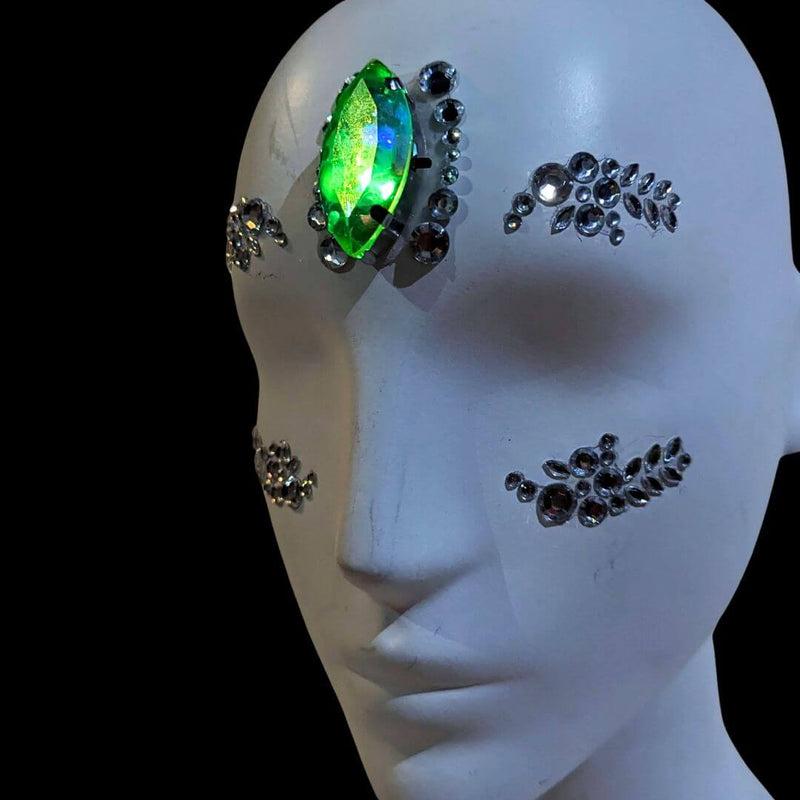 Mannequin face with rhinestone gem clusters and a green-glowing LED crystal on the brow, a color-changing LED rhinestone face sticker and face gems piece for festival makeup, alien looks, theatrical performances and avant-garde fashion events, Neon Cowboys