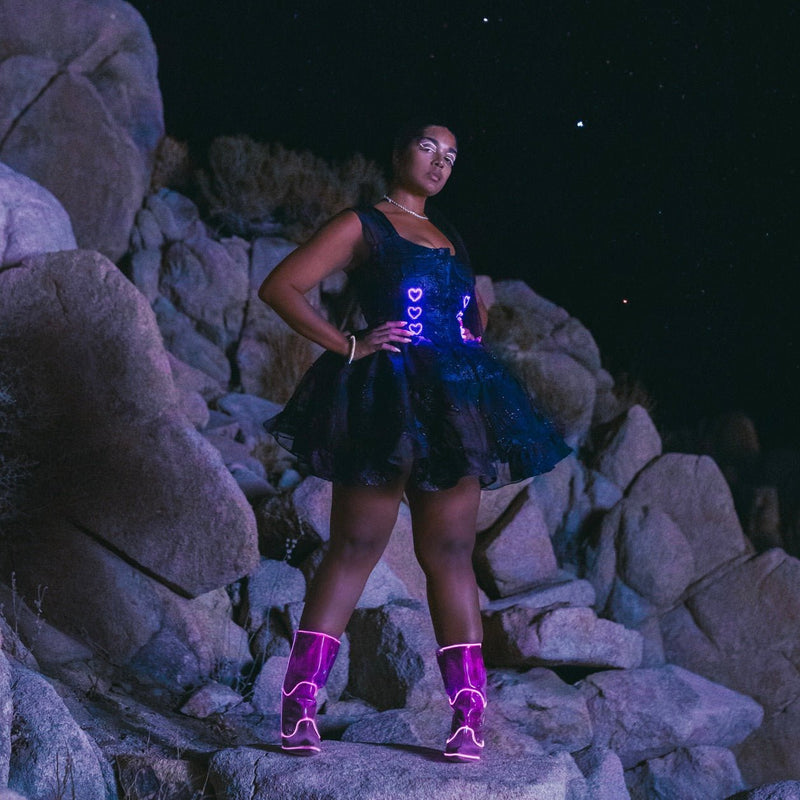 Model on desert rocks at night in a Neon Cowboys black ruffle dress glowing with pink LED hearts styled with boots, light up led dress festival stagewear for showgirls and cabaret performers, perfect for nightclub parties, Valentine's themed events and romantic date night outfits, Neon Cowboys