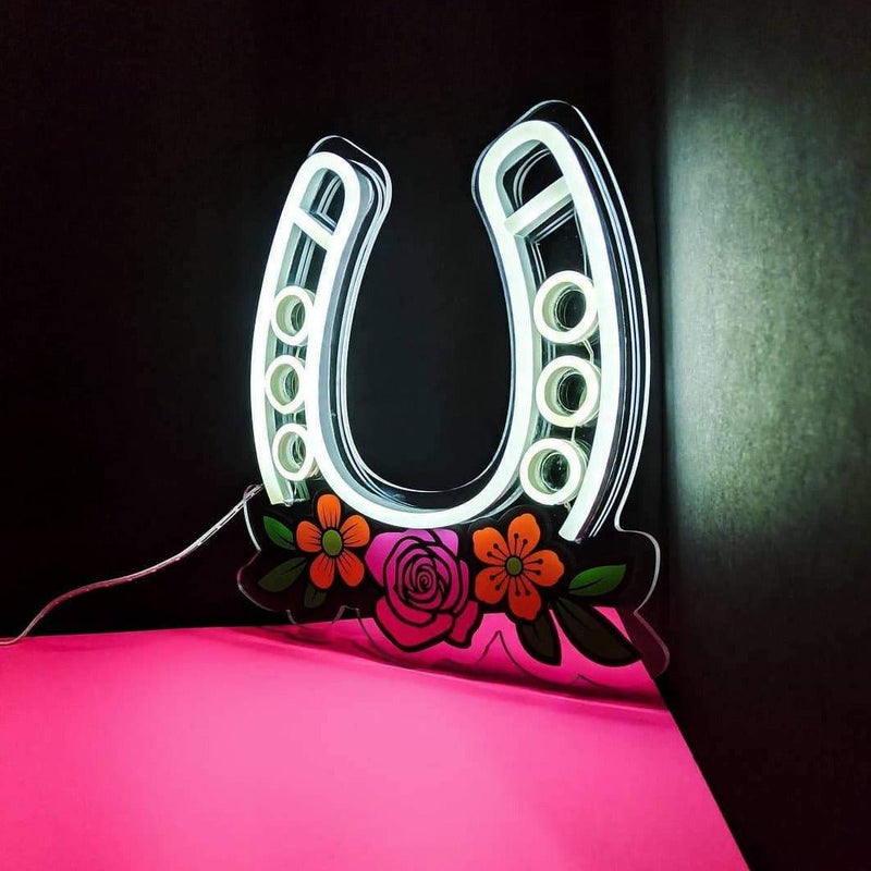 Good Luck horseshoe LED neon sign glowing white with painted roses on a pink surface, a good luck LED neon sign and glowing horseshoe wall light for western decor, bedroom and bar signage, home gallery walls and western-themed rooms, Neon Cowboys
